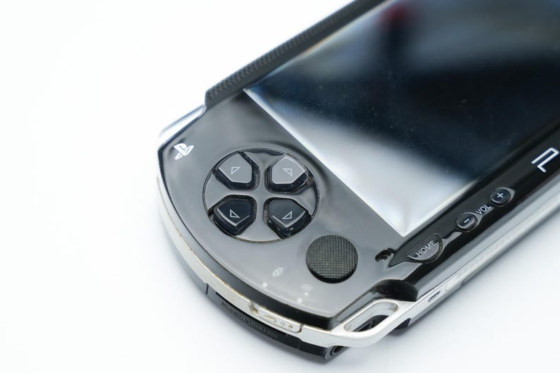 May include: A close-up of a black and silver handheld gaming console. The console features a directional pad with four buttons, a circular button, and a screen. The buttons are black, and the console has a silver trim.