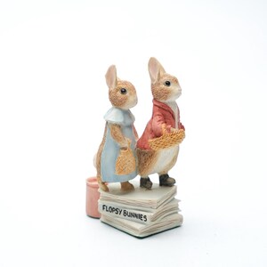 Puede incluir: Figura de dos conejos, uno con un vestido azul y otro con una chaqueta roja, de pie sobre una pila de libros con la inscripción "Flopsy Bunnies". Hay un pequeño cubo rosa junto a ellos. Los conejos son de color marrón claro y blanco.
