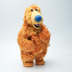 Op de afbeelding: Een pluche berenspeeltje met ruige oranje vacht, een lichtbruine snuit en een felblauwe neus. De beer heeft zwarte plastic ogen, een wijd open mond met een roze tong en staat op twee afgeronde voeten. De vacht heeft een lange, warrige textuur.