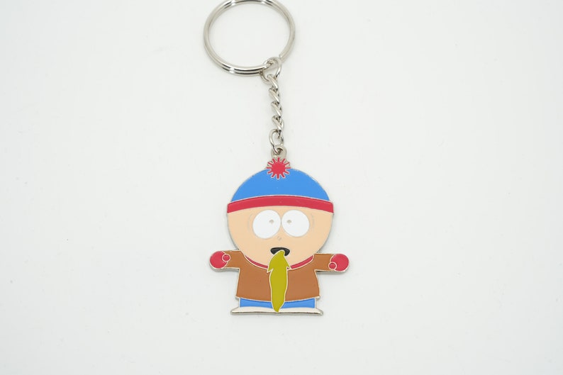 K&ouml;nnte beinhalten: Ein Metall-Schl&uuml;sselanh&auml;nger mit einer Cartoonfigur mit blauem Hut, rotem Rand und brauner Jacke. Die Figur hat einen &uuml;berraschten Gesichtsausdruck und eine gr&uuml;ne Substanz kommt aus ihrem Mund. Der Schl&uuml;sselanh&auml;nger hat eine silberne Kette und einen Ring.