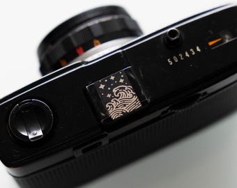 May include: A black 35mm film camera with a silver lens and a decorative sticker on the top. The sticker features a black and white design of waves and stars. The camera has a serial number "502434" printed on the top.