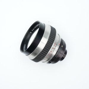 Può includere: Un obiettivo per fotocamera argentato e nero con una scala di profondità di campo contrassegnata in piedi e metri. L'obiettivo ha un anello di gomma nero attorno all'elemento anteriore.