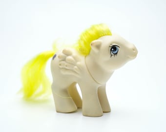 Può includere: Un giocattolo di pony bianco con i capelli gialli e un occhio blu. Il pony ha un'ala bianca e viola sulla schiena.
