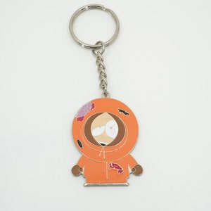 K&ouml;nnte beinhalten: Metall-Schl&uuml;sselanh&auml;nger mit einem Cartoon-Charakter in einem orangefarbenen Kapuzenparka. Der Schl&uuml;sselanh&auml;nger hat eine silberne Kette und einen Ring. Die Figur hat ein braunes Gesicht und tr&auml;gt eine orangefarbene Kapuze.