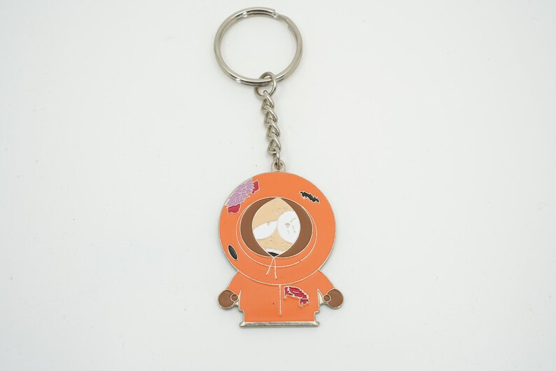 K&ouml;nnte beinhalten: Metall-Schl&uuml;sselanh&auml;nger mit einem Cartoon-Charakter in einem orangefarbenen Kapuzenparka. Der Schl&uuml;sselanh&auml;nger hat eine silberne Kette und einen Ring. Die Figur hat ein braunes Gesicht und tr&auml;gt eine orangefarbene Kapuze.