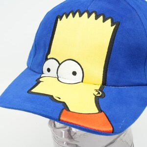 May include: A blue baseball cap featuring a yellow cartoon character of Bart Simpson from The Simpsons. The character's face is on the front of the cap.