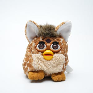 Op de afbeelding: Een bruin en bruin Furby-speelgoed met grote bruine ogen, een gele snavel en witte oren. De Furby heeft een textuurpatroon en een zachte, harige borst. Het is een retro elektronisch speelgoed.