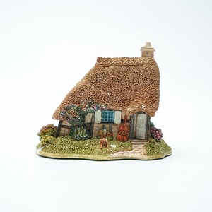 Op de afbeelding: Miniatuurbeeldje van een huisje met een rieten dak, een klein blauw raam en een deur. Het huisje is omgeven door groen en bloemen, met een viool die tegen de deur leunt. De figurine staat op een witte achtergrond.