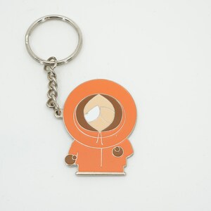 K&ouml;nnte beinhalten: Orangener und brauner Emaille-Schl&uuml;sselanh&auml;nger mit der Figur Kenny aus South Park. Der Schl&uuml;sselanh&auml;nger hat eine silberne Kette und einen Ring. Die Figur tr&auml;gt einen orangefarbenen Parka mit Kapuze.