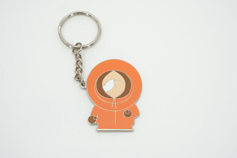 K&ouml;nnte beinhalten: Orangener und brauner Emaille-Schl&uuml;sselanh&auml;nger mit der Figur Kenny aus South Park. Der Schl&uuml;sselanh&auml;nger hat eine silberne Kette und einen Ring. Die Figur tr&auml;gt einen orangefarbenen Parka mit Kapuze.