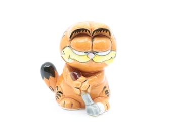 May include: A ceramic figurine of Garfield the cat holding a wrench. Garfield is orange with black stripes and a grumpy expression.