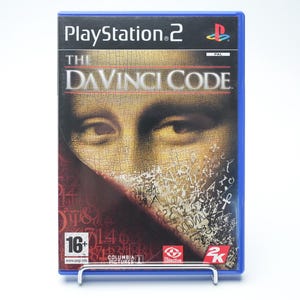 Puede incluir: Caja de juego de PlayStation 2 para "The Da Vinci Code". La portada presenta un primer plano de un rostro con superposición de texto. La caja es azul con el logotipo de PlayStation 2 en la parte superior. El juego está clasificado para mayores de 16 años.