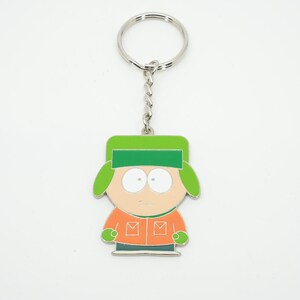 K&ouml;nnte beinhalten: Ein Schl&uuml;sselanh&auml;nger mit einer Cartoonfigur mit gr&uuml;nem Hut, orangefarbener Jacke und neutralem Gesichtsausdruck. Die Figur ist an einer silbernen Kette und einem Schl&uuml;sselring befestigt. Das Design ist schwarz umrandet.