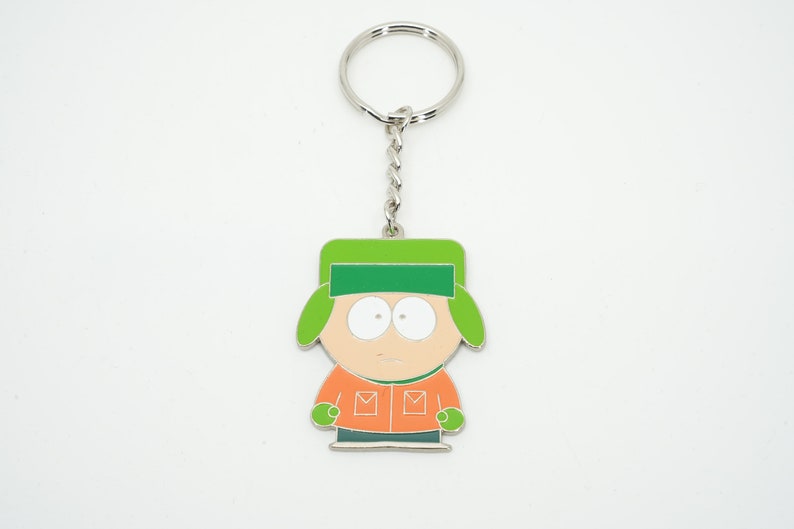 K&ouml;nnte beinhalten: Ein Schl&uuml;sselanh&auml;nger mit einer Cartoonfigur mit gr&uuml;nem Hut, orangefarbener Jacke und neutralem Gesichtsausdruck. Die Figur ist an einer silbernen Kette und einem Schl&uuml;sselring befestigt. Das Design ist schwarz umrandet.
