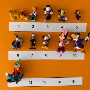 K&ouml;nnte beinhalten: Eine Sammlung von 12 kleinen Plastikfiguren von Disney-Charakteren, darunter Mickey Mouse, Minnie Mouse, Donald Duck, Goofy und Pluto. Die Figuren sind von 1 bis 12 nummeriert und in zwei Reihen auf einem wei&szlig;en Hintergrund angeordnet.