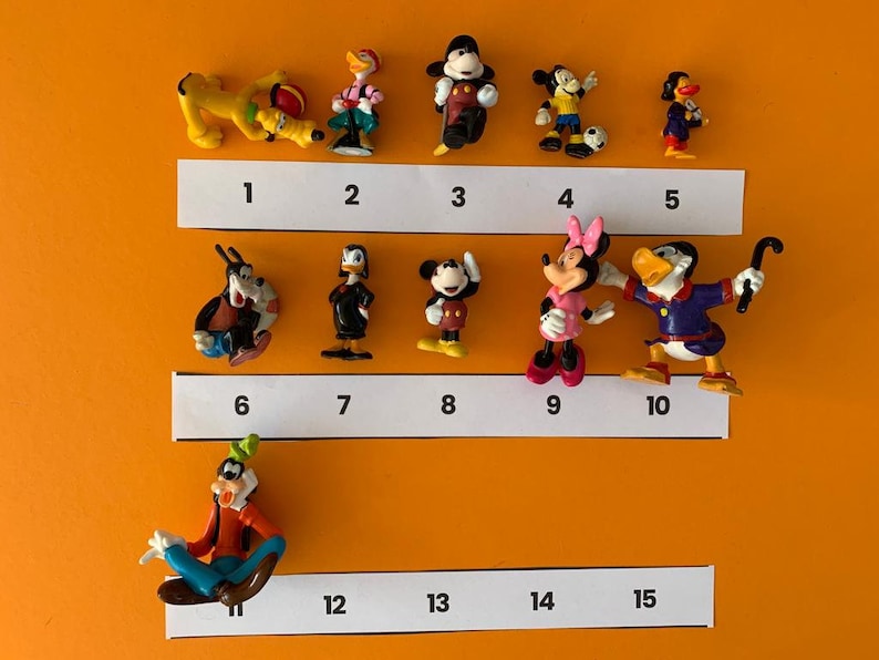 K&ouml;nnte beinhalten: Eine Sammlung von 12 kleinen Plastikfiguren von Disney-Charakteren, darunter Mickey Mouse, Minnie Mouse, Donald Duck, Goofy und Pluto. Die Figuren sind von 1 bis 12 nummeriert und in zwei Reihen auf einem wei&szlig;en Hintergrund angeordnet.