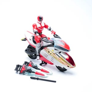 Könnte beinhalten: Rote und weiße Actionfigur auf einem passenden Motorrad. Die Figur trägt einen roten Anzug und Helm. Das Motorrad hat goldene und silberne Akzente. Zusätzliches Zubehör ist enthalten.