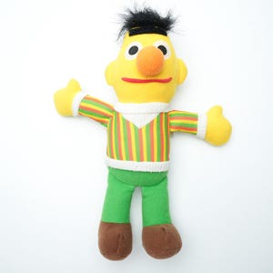 May include: A plush toy of a yellow character with a large orange nose, black hair, and a striped shirt with green pants. The character has its arms outstretched and brown shoes. The toy is on a white background.