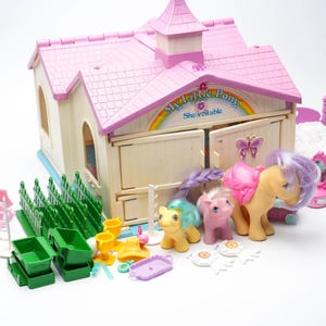 Może przedstawiać: Zestaw do zabawy My Little Pony Show Stable w stylu vintage. Zestaw zawiera stajnię z różowym dachem, trzy zabawki kucyki i różne akcesoria, takie jak ogrodzenia, wiadra i łóżko. Na stajni widnieje napis "My Little Pony Show Stable".