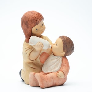 Op de afbeelding: Een keramieken beeldje dat een persoon voorstelt die een baby een flesje geeft. De persoon heeft bruin haar en draagt een beige outfit. De baby, in een perzikkleurige outfit, zit en draagt een witte slab. Het beeldje staat op een witte achtergrond.