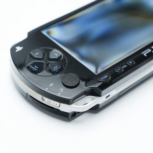 May include: Black PSP handheld game console with a silver trim. The console features a directional pad, action buttons, and a screen. The PSP logo is visible on the top right. The device is on a white background.