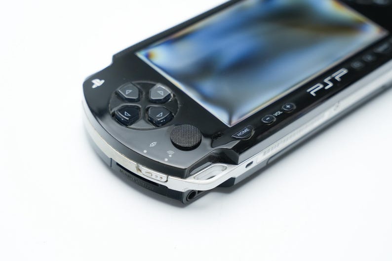 May include: Black PSP handheld game console with a silver trim. The console features a directional pad, action buttons, and a screen. The PSP logo is visible on the top right. The device is on a white background.