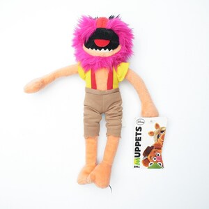 Puede incluir: Un peluche del personaje Animal de los Muppets. El juguete tiene una cabeza rosa y peluda, una boca negra abierta y brazos y piernas naranjas. Lleva una camisa amarilla con tirantes rojos y pantalones marrones. Una etiqueta con el logo de los Muppets está adjunta.