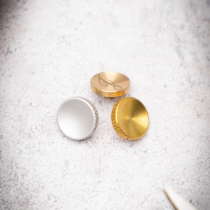 Puede incluir: Tres botones redondos para reloj, uno plateado, uno dorado y uno dorado con un diseño. Los botones están sobre una superficie blanca.