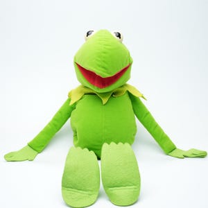 Könnte beinhalten: Ein Plüschtier von Kermit dem Frosch, mit einem hellgrünen Körper, Armen und Beinen. Der Frosch hat einen roten Mund, schwarze Augen und einen gelben Kragen. Das Spielzeug sitzt mit ausgestreckten Armen und Beinen.