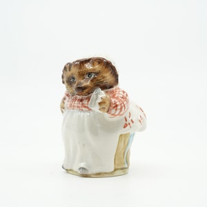 May include: A ceramic figurine of a hedgehog wearing a red and white gingham dress and a white apron. The hedgehog is holding a small white object in its right paw.