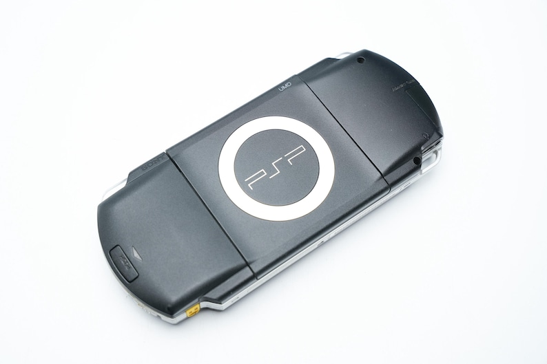 May include: A black handheld PSP game console with a white PSP logo in the center. The console is angled slightly, showing the top and front. The background is plain white.