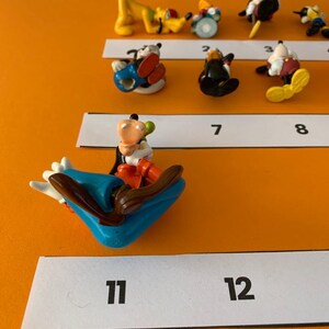 K&ouml;nnte beinhalten: Eine Sammlung von 13 bunten Plastikfiguren von Disney-Charakteren, darunter Mickey Mouse, Minnie Mouse, Goofy und Donald Duck. Die Figuren sind in einer Reihe auf einer wei&szlig;en Oberfl&auml;che mit den Zahlen 1 bis 13 angeordnet.