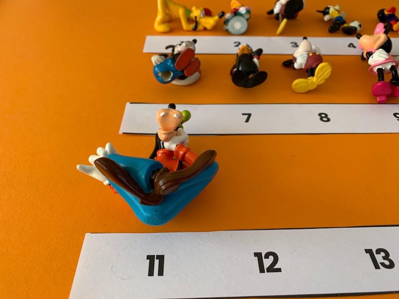 K&ouml;nnte beinhalten: Eine Sammlung von 13 bunten Plastikfiguren von Disney-Charakteren, darunter Mickey Mouse, Minnie Mouse, Goofy und Donald Duck. Die Figuren sind in einer Reihe auf einer wei&szlig;en Oberfl&auml;che mit den Zahlen 1 bis 13 angeordnet.