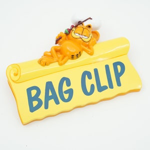 Puede incluir: Un clip de bolsa de plástico amarillo con un gato naranja de dibujos animados que lleva un gorro de cocinero y sostiene un pincho. El texto "BAG CLIP" está impreso en azul en el clip.