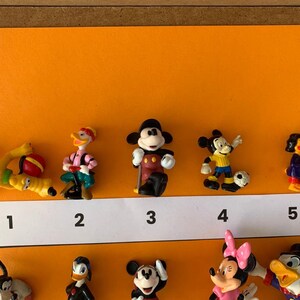 K&ouml;nnte beinhalten: Ein Set mit zehn kleinen Plastikfiguren von Disney-Charakteren, darunter Mickey Mouse, Donald Duck, Goofy und Pluto. Die Figuren sind von 1 bis 10 nummeriert und in einer Reihe auf einer wei&szlig;en Oberfl&auml;che angeordnet.