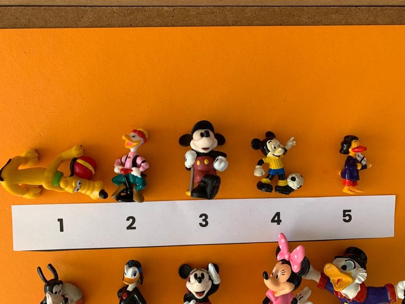 K&ouml;nnte beinhalten: Ein Set mit zehn kleinen Plastikfiguren von Disney-Charakteren, darunter Mickey Mouse, Donald Duck, Goofy und Pluto. Die Figuren sind von 1 bis 10 nummeriert und in einer Reihe auf einer wei&szlig;en Oberfl&auml;che angeordnet.