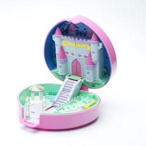 Op de afbeelding: Een vintage hartvormige speelset, open om een miniatuurkasteel met roze torentjes en een roze deur te onthullen. De basis heeft een tuin met een prieel en een klein zwembad. Het speelgoed is voornamelijk roze, blauw en wit.
