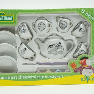 Op de afbeelding: Een Sesame Street-thema theeservies voor kinderen, met een theepot, kopjes, schoteltjes en een kannetje. De set is wit met zwarte Sesame Street-personage illustraties en wordt geleverd met verf om te decoreren.