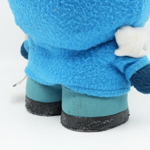 Puede incluir: Un juguete de peluche azul con una cara blanca y zapatos negros. El juguete lleva una chaqueta azul con capucha.
