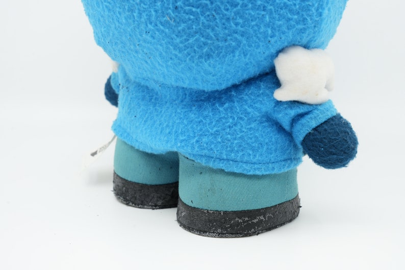 Puede incluir: Un juguete de peluche azul con una cara blanca y zapatos negros. El juguete lleva una chaqueta azul con capucha.