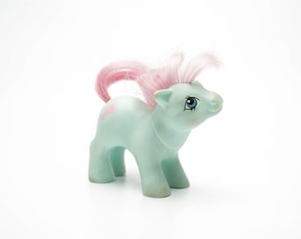 Può includere: Un giocattolo di pony di plastica blu chiaro con i capelli rosa e un occhio blu. Il pony ha un cuore rosa su un lato.