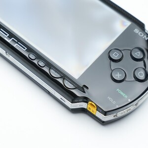 May include: Black Sony PSP handheld game console. The device has a glossy black finish with silver trim. Visible buttons include directional buttons, action buttons, and power and hold switches. The screen is visible.