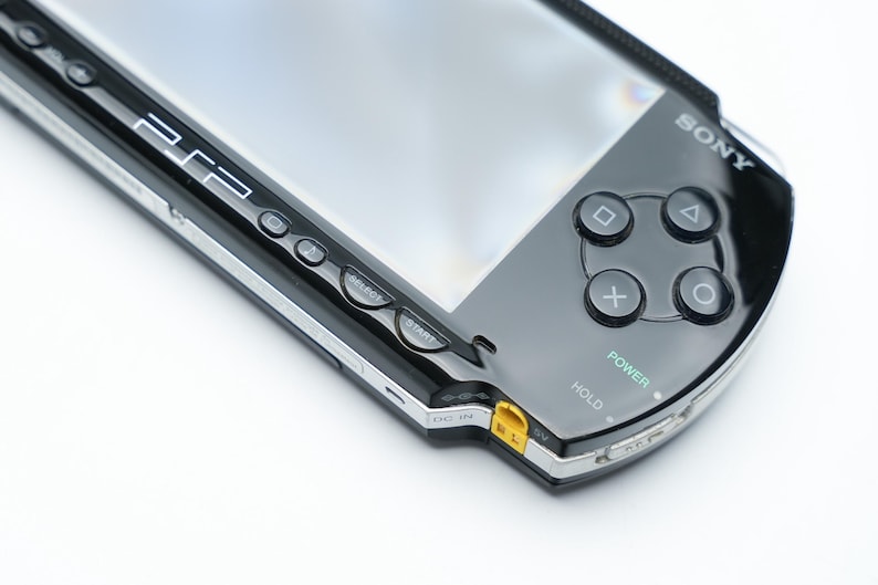 May include: Black Sony PSP handheld game console. The device has a glossy black finish with silver trim. Visible buttons include directional buttons, action buttons, and power and hold switches. The screen is visible.