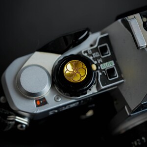 May include: Close-up of a silver camera with a black dial and a gold button with a flower design. The button is labeled "AUTO".