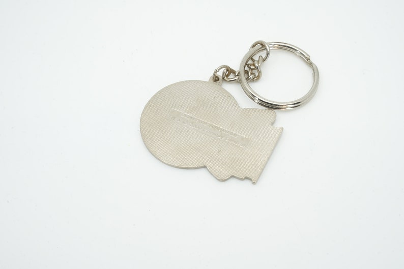 K&ouml;nnte beinhalten: Silberfarbener Metall-Schl&uuml;sselanh&auml;nger mit kreisf&ouml;rmigem und geometrischem Design. Der Schl&uuml;sselanh&auml;nger hat einen geteilten Ring f&uuml;r Schl&uuml;ssel und einen kleinen rechteckigen Text. Das Design befindet sich auf wei&szlig;em Hintergrund.