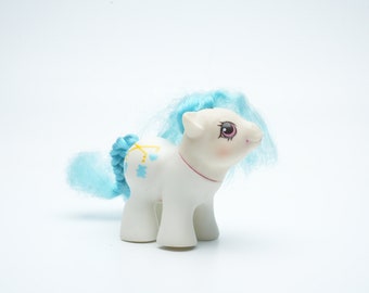 Puede incluir: Un juguete de pony blanco con pelo azul y un patrón de estrellas azules, amarillas y blancas en su costado. El pony tiene ojos rosas y una nariz rosa.