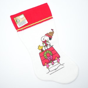 以下が含まれることがあります： 赤色のカフと金色のトリムが付いた白いクリスマスストッキング。ストッキングには、緑色の花輪とピンクのリボンが付いた赤い犬小屋にいるスヌーピーとウッドストックのグラフィックが描かれています。タグがカフに付いています。