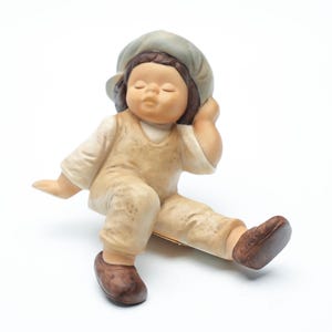 Könnte beinhalten: Keramikfigur eines sitzenden Kindes mit geschlossenen Augen, das ein beiges Outfit und eine graue Mütze trägt. Die Figur hat braune Schuhe und stützt ihren Kopf auf ihre Hand. Die Farbpalette ist gedämpft, mit Fokus auf Beige- und Brauntönen.