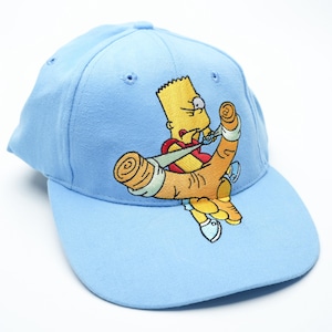 May include: Light blue baseball cap featuring an embroidered image of Bart Simpson from The Simpsons cartoon. Bart is depicted holding a slingshot. The cap has a curved brim and a six-panel design, suitable for casual wear.