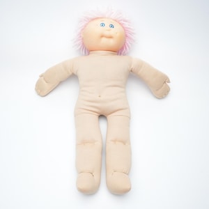 Może przedstawiać: Miękka, beżowa lalka z okrągłą głową, niebieskimi oczami i różowymi włosami. Lalka ma materiałowy korpus z wyciągniętymi ramionami i nogami. Lalka jest na białym tle.
